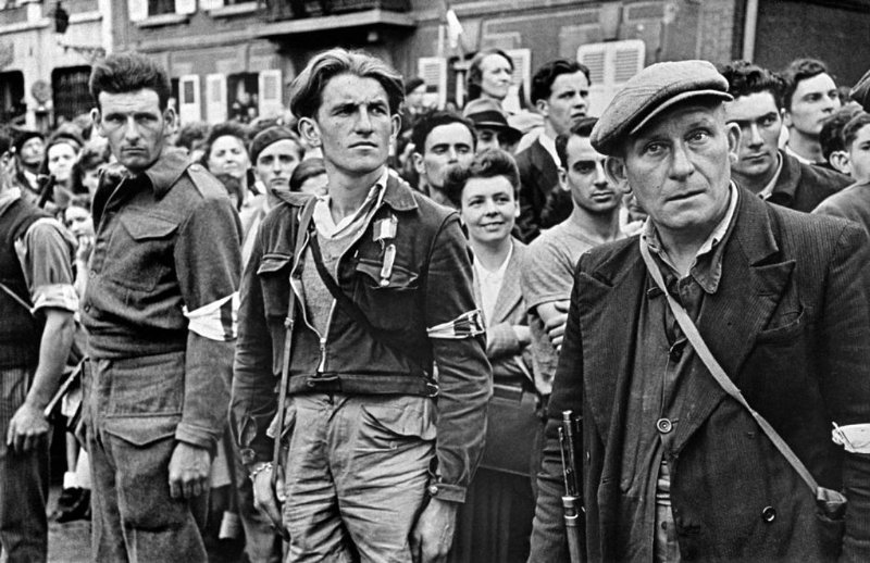 Des résistants français se rangent dans les rues de Chartres pour écouter le discours du chef des Forces françaises libres, le général Charles de Gaulle, proclamant la libération complète de la ville de l’occupation allemande. Chartres a été libérée le 18 août 1944 par la 5e division d’infanterie et la 7e division blindée de l’armée américaine, appartenant au XXe corps de la 3e armée américaine (United States Army Central). Chartres, Eure-et-Loir, Région Centre, France. 23 août 1944. Photo prise par Robert Capa.<br /><br />Source: https://bag-of-dirt.tumblr.com/post/92479581260/french-resistance-fighters-line-the-streets-of